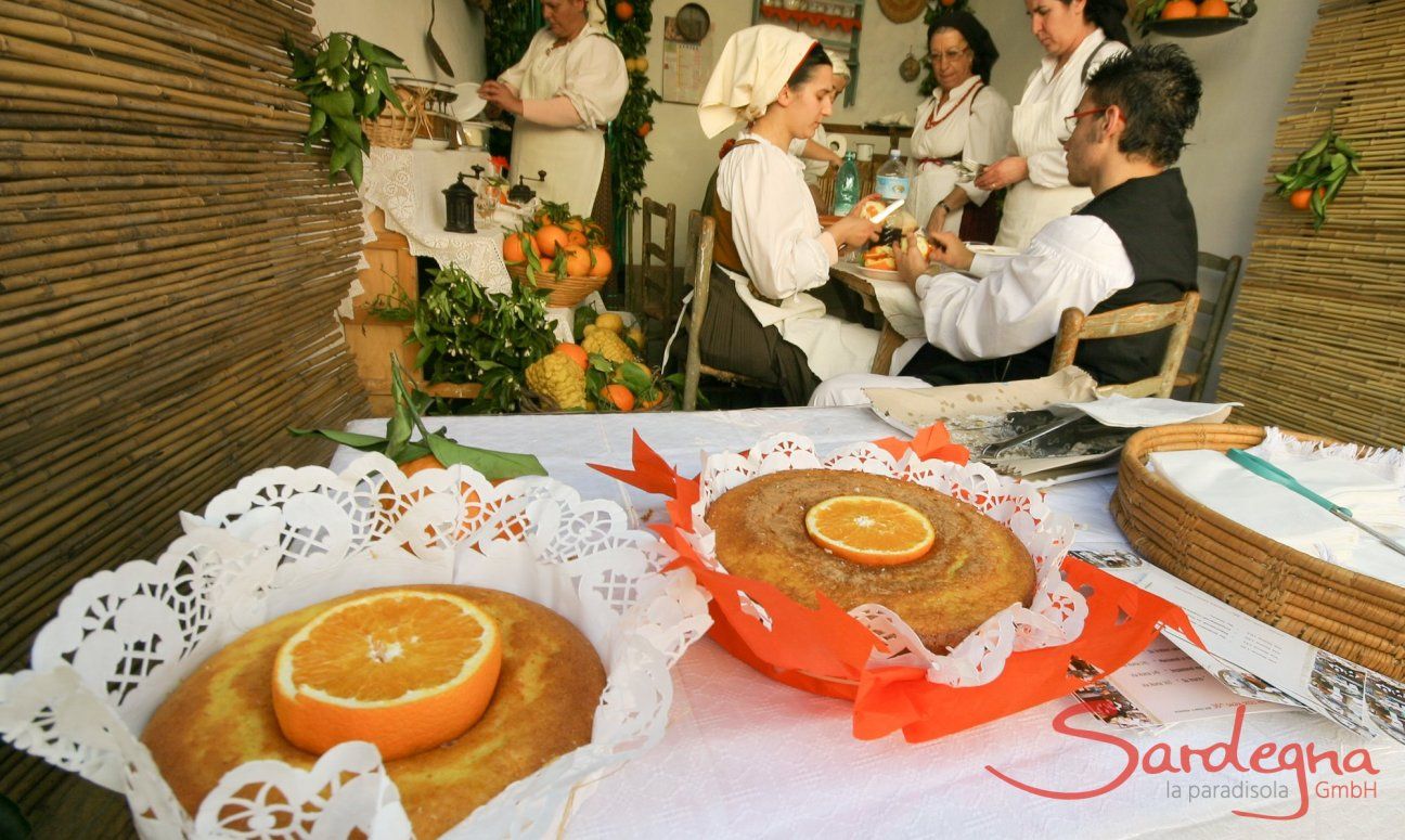 Präsentation von Orangenkuchen beim Tag der offenen Tür, im Zimmer dahinter Sarden in Trachten bei der Zubereitung der Kuchen