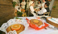 Präsentation von Orangenkuchen beim Tag der offenen Tür, im Zimmer dahinter Sarden in Trachten bei der Zubereitung der Kuchen