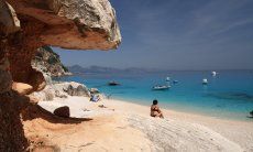 Frau sitzt am Strand vor den Felsen von Cala Luna und schaut auf die Motorboote auf dem Meer