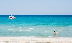 Frau läuft am weißen Strand von La Cinta vor dem hellblauen Meer mit einem weiß und roten Boot